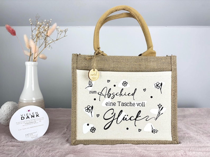 Pu&ograve; includere: Una borsa tote in iuta con un pannello frontale color crema con il testo tedesco "zum Abschied eine Tasche voll Gl&uuml;ck" in scrittura nera, che significa "una borsa piena di fortuna". Ha un manico in iuta e una piccola etichetta di legno.