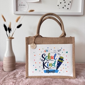 May include: A rectangular jute tote bag with two handles and a white front panel. The panel features colourful confetti dots, the text "ABC", "Schul Kind", and "Bent", along with a graphic of a blue cone-shaped gift bag with white stars.