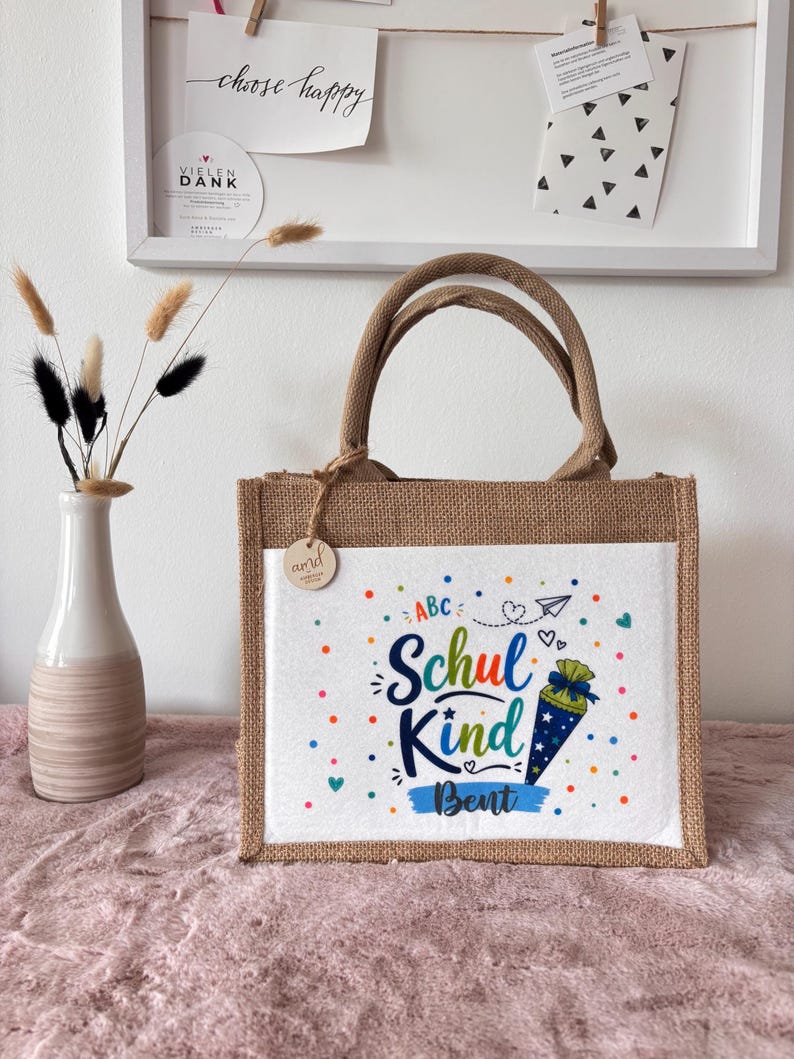 May include: A rectangular jute tote bag with two handles and a white front panel. The panel features colourful confetti dots, the text "ABC", "Schul Kind", and "Bent", along with a graphic of a blue cone-shaped gift bag with white stars.