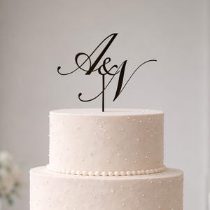 May include: A three-tiered white cake decorated with small white dots and a beaded border on each tier. A black cake topper featuring the stylised letters A and N connected by an ampersand sits on the top tier of the cake.