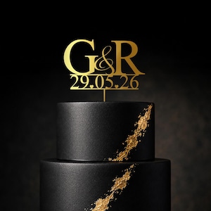 May include: A three-tiered black cake with gold detailing. The cake is decorated with a gold cake topper displaying "G&R" and the date "29.05.26". The cake has a contemporary design, suitable for a wedding or special occasion.
