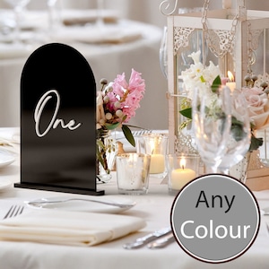 May include: Black arch-shaped table number with the word "One" written in white. The table number is on a white tablecloth with candles, flowers, and glasses.