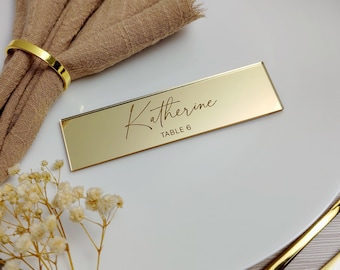 May include: A gold rectangular place card with the name "Katherine" and "TABLE 6" in elegant script. The card sits on a white plate, next to a gold napkin ring and a folded tan napkin. Floral accents are visible.