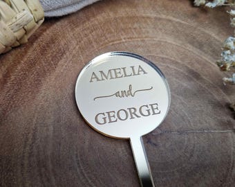 May include: A round, silver, mirrored sign with the text "Amelia and George" engraved on it. The sign is attached to a silver stick.