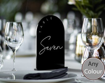 Pode incluir: Número de mesa de acrílico preto com forma de arco e o texto "TABLE" e "Seven" em branco. O número de mesa está sobre uma mesa branca com uma toalha de mesa preta e um copo de água.