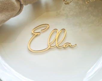 May include: A gold-colored acrylic nameplate with the name "Ella" in a cursive font. The nameplate is placed on a white plate. The image is well-lit, highlighting the nameplate's elegant design.
