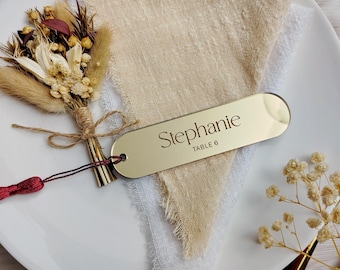 May include: A gold-colored place card with the name "Stephanie" and "TABLE 6" printed on it, resting on a white plate with a beige linen napkin. A small bouquet of dried flowers and a red tassel are also present.