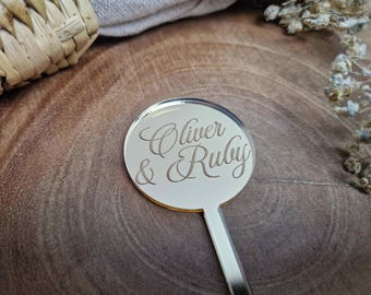 May include: A round, mirrored acrylic wedding favor with the names "Oliver & Ruby" engraved in elegant script. The favor has a small handle and is set against a wooden surface.
