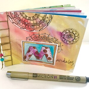 Puede incluir: Un diario de acuarela con un diseño de mandala dibujado a mano en la portada. El diario está encuadernado con hilo y tiene una portada de papel marrón. La palabra "mandalas" está escrita en la portada con tinta negra. También se muestra una pluma de tinta de archivo Micron 01.