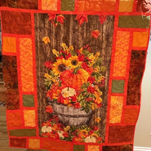 May include: A handmade autumn-themed quilt featuring a central panel with a rustic wooden background and a still life of pumpkins, sunflowers, and fall foliage in a metal bucket. The quilt is bordered with orange, brown, and green patchwork.