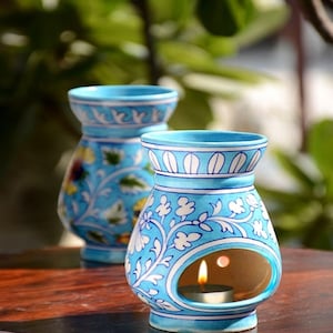 Può includere: Due scaldatori di olio in ceramica blu con disegni floreali. Uno scaldatore ha una candela accesa all'interno, visibile attraverso un'apertura rotonda. Gli scaldatori sono decorati con motivi floreali bianchi e gialli e poggiano su una superficie di legno.