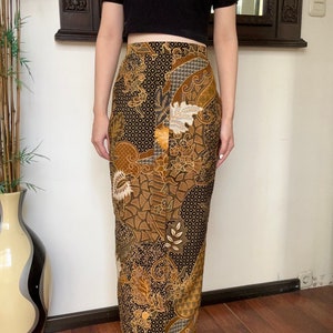 Op de afbeelding: Een lange, zwarte rok met een bruine en gouden batikprint. De rok heeft een hoge taille en een aansluitende pasvorm.