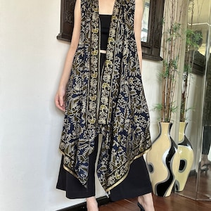 Op de afbeelding: Een lange, mouwloze, zwart-gouden geprinte vest met een gedrapeerd ontwerp. De vest heeft een V-hals en een gouden rand. De vest wordt gedragen over een zwart topje en een zwarte broek.