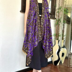 May include: A purple and gold patterned sleeveless vest with a draped, asymmetrical hem. The vest is made of a silky fabric and has a relaxed fit.