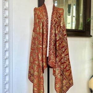 May include: A red and gold sleeveless vest with a paisley pattern. The vest has a long, flowing design and is made of a soft, silky fabric.