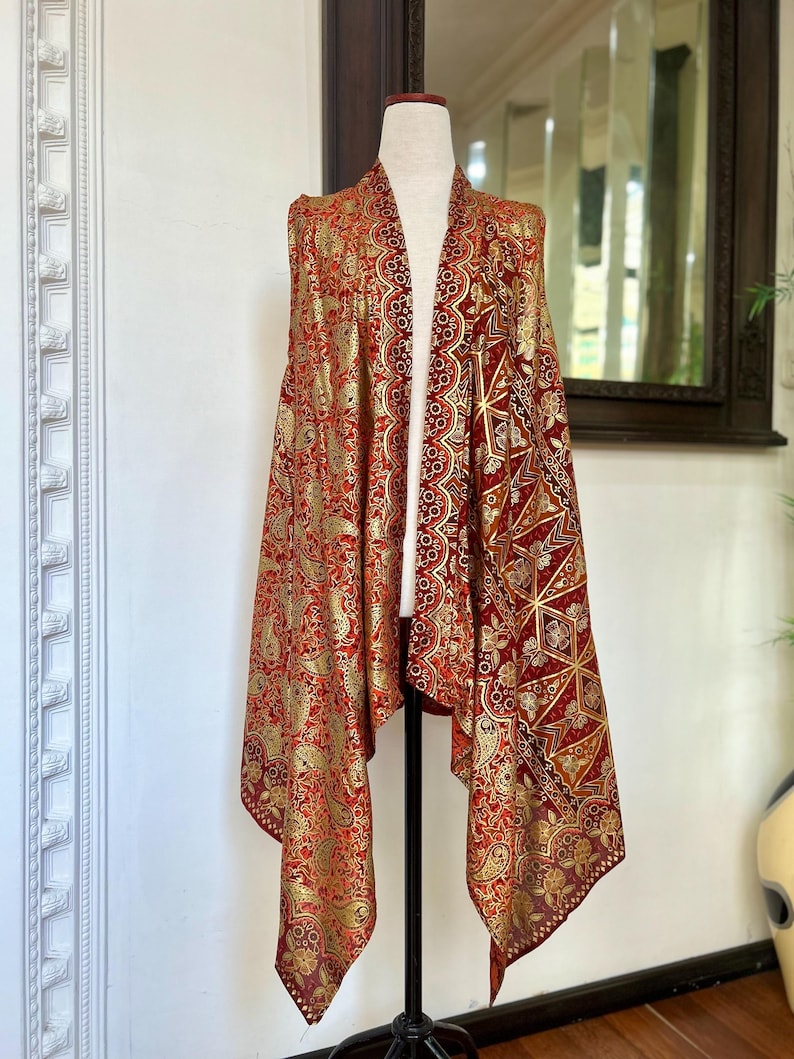May include: A red and gold sleeveless vest with a paisley pattern. The vest has a long, flowing design and is made of a soft, silky fabric.