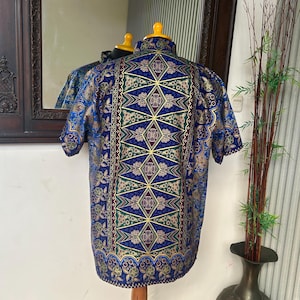 Op de afbeelding: Een overhemd met korte mouwen met een levendige blauwe achtergrond en een gedetailleerd geometrisch patroon in groen, goud en bordeauxrood. Het overhemd wordt getoond op een houten mannequin, die het ingewikkelde ontwerp laat zien.