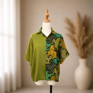 May include: A short-sleeved button-down shirt featuring a split design. The left side is solid olive green, while the right side displays a complex, multi-coloured batik pattern in shades of mustard yellow, teal, black, and green with geometric motifs.