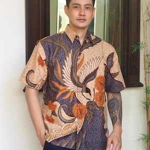 Op de afbeelding: Een beige shirt met korte mouwen met een kleurrijk batikpatroon met een vogel en bloemen. Het shirt heeft een knoopsluiting.