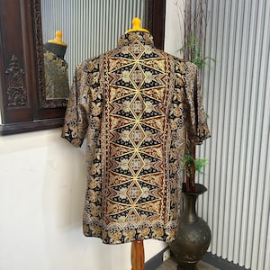 Op de afbeelding: Korte mouwen shirt met een gedetailleerd batikpatroon in zwart, bruin en goud. Het shirt wordt getoond op een houten mannequin, met de ingewikkelde geometrische en bloemmotieven. De kraag is geel.