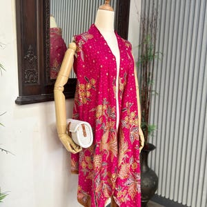 Può includere: Un gilet rosa acceso con apertura frontale e un motivo floreale e di uccelli in oro e marrone. Il gilet è esposto su un manichino, abbinato a una pochette bianca intrecciata. Il design del gilet presenta una stampa tradizionale dettagliata.