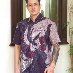 Op de afbeelding: Een paars en roze shirt met korte mouwen met een batikpatroon met een vogel en bloemen. Het shirt heeft een knoopsluiting en een ontspannen pasvorm.