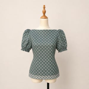Könnte beinhalten: Eine dunkelgrüne Bluse mit einem sich wiederholenden weißen Blumenmuster, einem weiten U-Boot-Ausschnitt und kurzen Puffärmeln mit gerafften elastischen Bündchen. Der Saum ist mit einem markanten weißen geometrischen Bordürenmuster versehen.