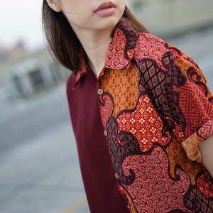 Può includere: Una camicia con colletto abbottonato a fantasia rosso e marrone. La camicia presenta un motivo geometrico con volute e quadrati.