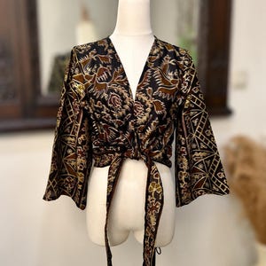 Könnte beinhalten: Ein schwarzes Wickeloberteil mit braunem und goldenem floralem Batikmuster, tiefem V-Ausschnitt und weiten Dreiviertelärmeln. Das Kleidungsstück verfügt über lange Stoffbänder an der vorderen Taille, die geknotet sind, um den Wickelverschluss zu sichern.