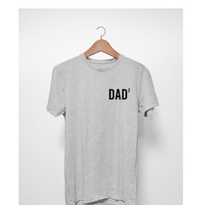 May include: A heather gray t-shirt with the text "DAD²" printed in black. The short-sleeved shirt is hanging on a wooden hanger against a white background. The text is centered on the chest.
