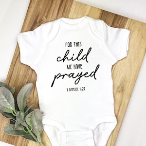Può includere: Body da bambino bianco con la scritta "FOR THIS child WE HAVE prayed 1 Samuel 1:27" in corsivo nero. Il body è appoggiato su un tagliere di legno con un rametto di verde.