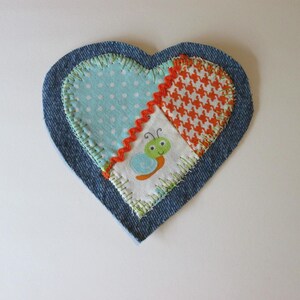 May include: A heart-shaped fabric patch featuring a dark blue denim border. The center is composed of three fabric sections: light blue with white polka dots, orange and white houndstooth, and white with a green and blue snail illustration.