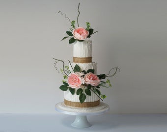 May include: A three-tiered white wedding cake decorated with burlap ribbon and pink flowers. The cake is on a white cake stand.