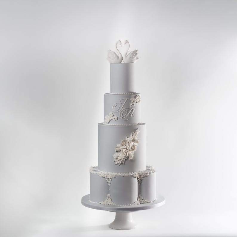 Puede incluir: Una tarta de boda gris claro de cinco pisos con decoraciones florales blancas y un par de figuras de cisnes en la parte superior. La tarta se exhibe sobre un pedestal blanco y presenta las iniciales "I A".