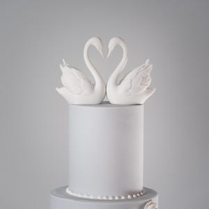 Puede incluir: Tarta de dos pisos gris claro, coronada con figuras de cisnes blancos que forman un coraz&oacute;n. La tarta tiene un dise&ntilde;o sencillo con un borde de perlas y un adorno floral.