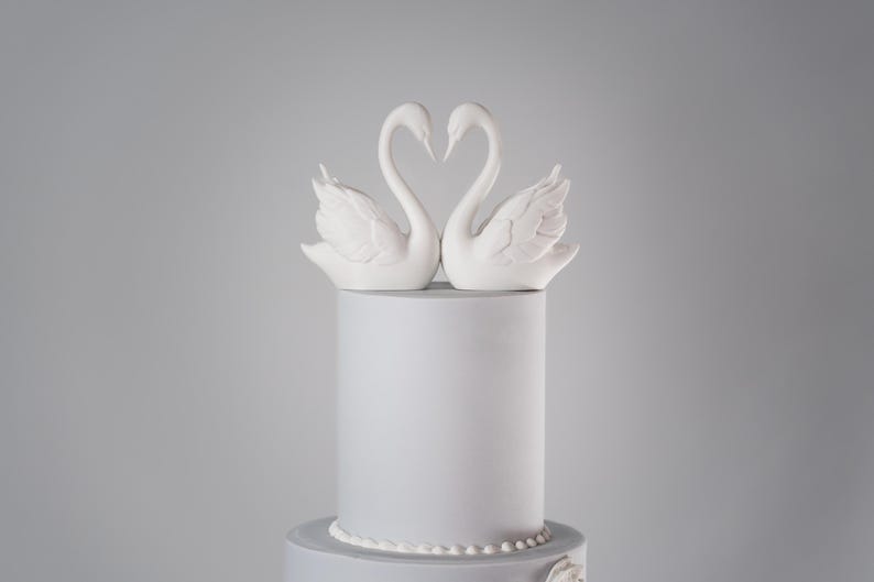 Puede incluir: Tarta de dos pisos gris claro, coronada con figuras de cisnes blancos que forman un coraz&oacute;n. La tarta tiene un dise&ntilde;o sencillo con un borde de perlas y un adorno floral.