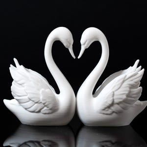 Puede incluir: Dos figuras de cisnes blancos enfrentadas, con sus cuellos formando un coraz&oacute;n. Los cisnes tienen texturas de plumas detalladas y est&aacute;n sobre un fondo negro, creando una exhibici&oacute;n rom&aacute;ntica y elegante.
