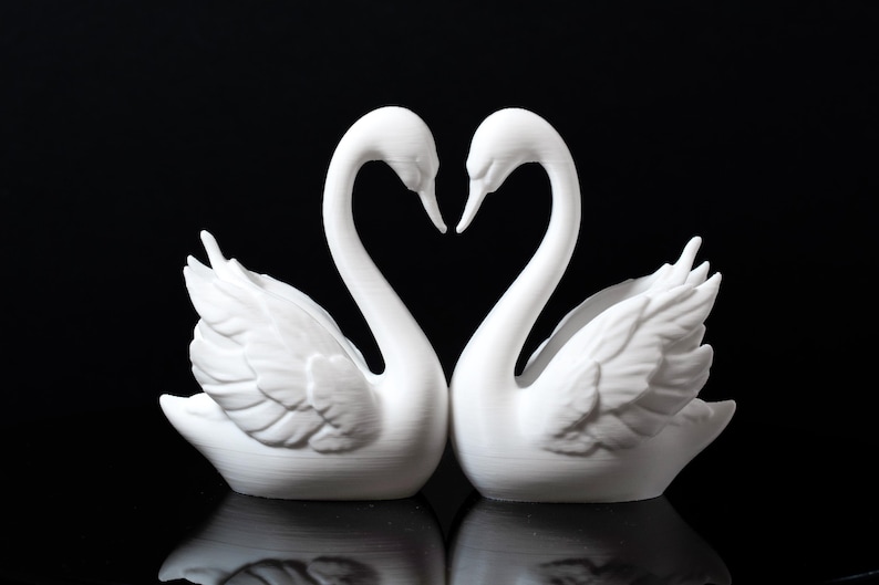 Puede incluir: Dos figuras de cisnes blancos enfrentadas, con sus cuellos formando un coraz&oacute;n. Los cisnes tienen texturas de plumas detalladas y est&aacute;n sobre un fondo negro, creando una exhibici&oacute;n rom&aacute;ntica y elegante.