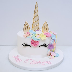 Puede incluir: Pastel de unicornio blanco con un cuerno y orejas de madera. El pastel está decorado con flores de glaseado rosa, amarillo y azul y las palabras "Happy 5th Birthday" en glaseado rosa.