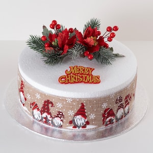 May include: A round Christmas cake with white icing, decorated with red flowers, berries, and evergreen sprigs. The cake has a ribbon with gnome illustrations and the words "MERRY CHRISTMAS".