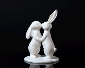 Puede incluir: Escultura blanca de dos conejos besándose, tomados de la mano. Los conejos están sobre una base redonda. El fondo es negro.
