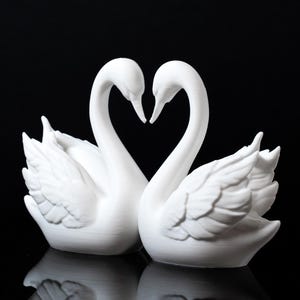 Puede incluir: Dos figuras de cisnes blancos enfrentadas, con sus cuellos formando un coraz&oacute;n. Los cisnes tienen texturas de plumas detalladas y est&aacute;n sobre un fondo negro, creando una exhibici&oacute;n rom&aacute;ntica y elegante.