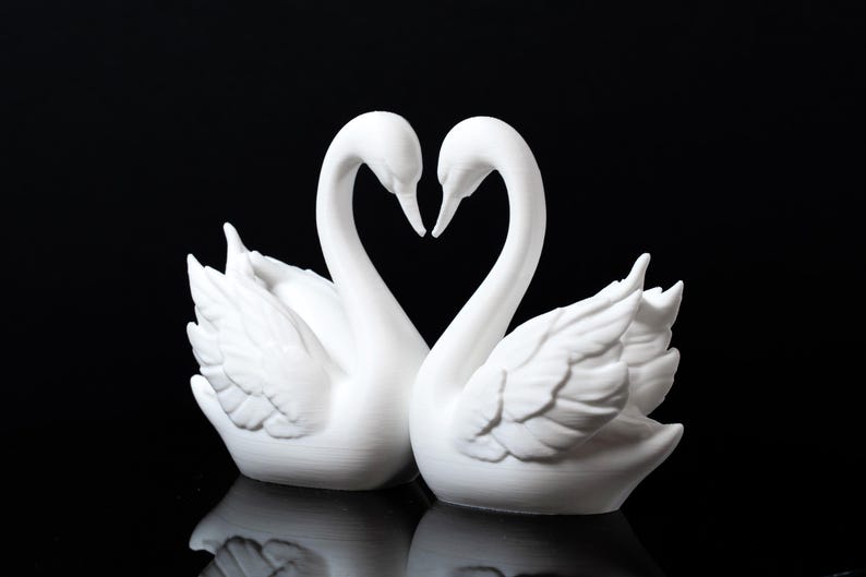 Puede incluir: Dos figuras de cisnes blancos enfrentadas, con sus cuellos formando un coraz&oacute;n. Los cisnes tienen texturas de plumas detalladas y est&aacute;n sobre un fondo negro, creando una exhibici&oacute;n rom&aacute;ntica y elegante.