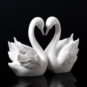 Puede incluir: Dos figuras de cisnes blancos, con los cuellos entrelazados formando un coraz&oacute;n. Los cisnes est&aacute;n juntos, con detalles de plumas. Las esculturas est&aacute;n sobre un fondo negro, pareciendo hechas de un material mate.