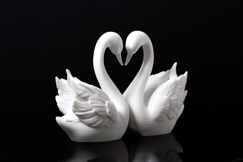 Puede incluir: Dos figuras de cisnes blancos, con los cuellos entrelazados formando un coraz&oacute;n. Los cisnes est&aacute;n juntos, con detalles de plumas. Las esculturas est&aacute;n sobre un fondo negro, pareciendo hechas de un material mate.