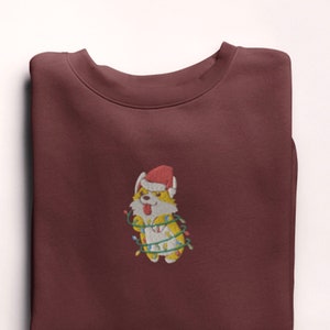 Może przedstawiać: Bordowa bluza z haftowanym obrazem corgi w czapce Świętego Mikołaja, zaplątanego w świąteczne lampki.