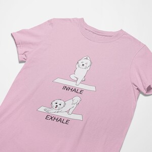May include: Pink t-shirt with a white dog doing yoga poses. The dog is in a downward dog pose with the text "Exhale" and in a standing pose with the text "Inhale".