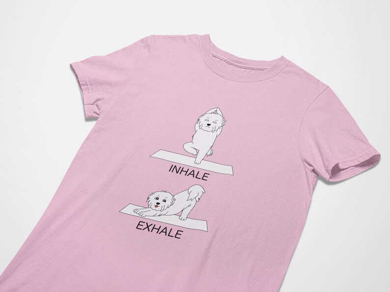 May include: Pink t-shirt with a white dog doing yoga poses. The dog is in a downward dog pose with the text "Exhale" and in a standing pose with the text "Inhale".