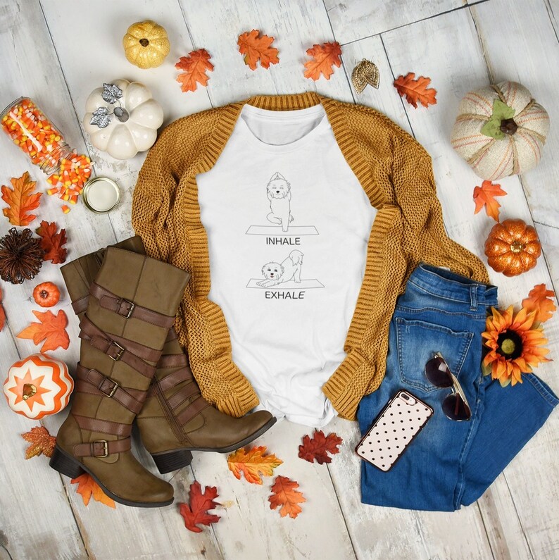 May include: White t-shirt with a graphic of a dog doing yoga poses. The text on the shirt says "inhale" and "exhale". The shirt is paired with a brown cardigan, blue jeans, brown boots, and fall-themed decorations.