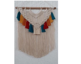 Pode incluir: Uma tapeçaria de macramé de parede com uma barra de madeira na parte superior. O macramé é feito com corda de cor natural e tem uma franja na parte inferior. O macramé tem um design geométrico com borlas coloridas em tons de laranja, vermelho, azul e verde.